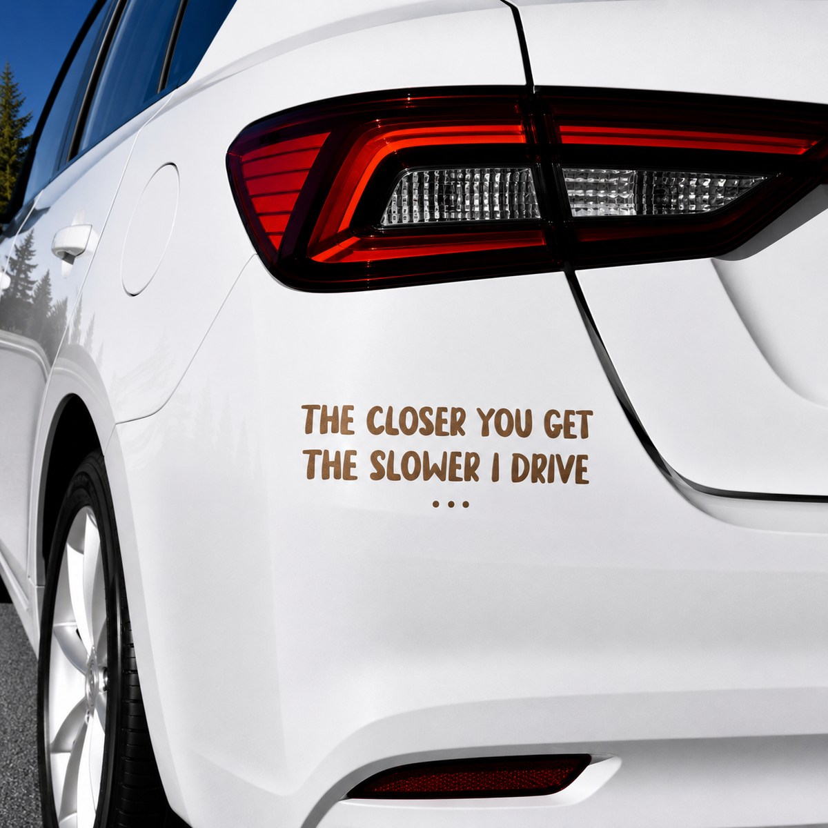 A white car rear quarter view with a brown text decal reading 'THE CLOSER YOU GET THE SLOWER I DRIVE' applied near the left side of the trunk.