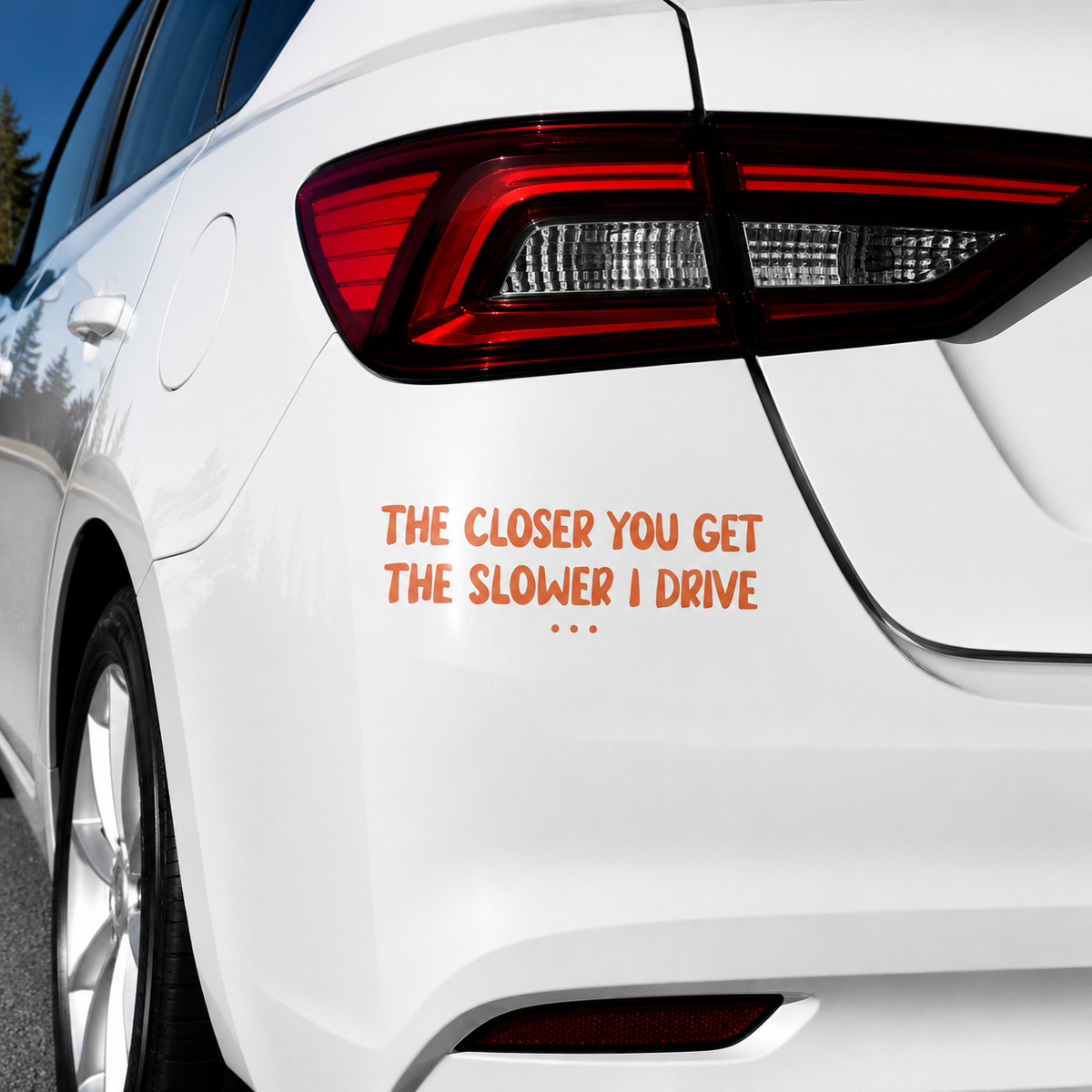 A white car rear quarter view with orange text decal reading 'THE CLOSER YOU GET THE SLOWER I DRIVE' applied near the lower right side of the trunk.