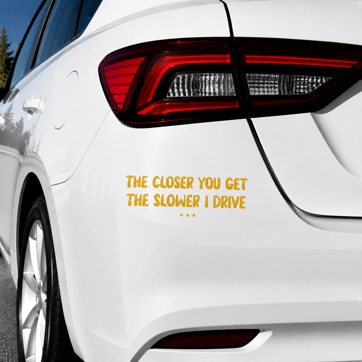 A yellow vinyl decal sticker applied to the rear left side of a white car, featuring the humorous text 'THE CLOSER YOU GET THE SLOWER I DRIVE .