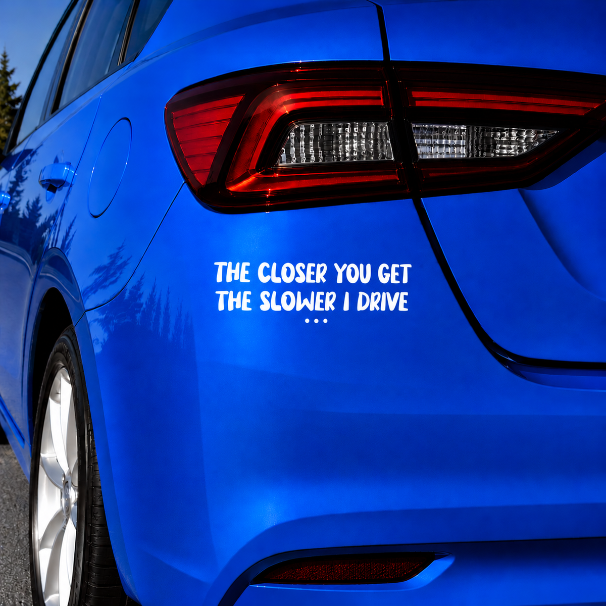 A bright blue car rear quarter view with white text decal reading 'THE CLOSER YOU GET THE SLOWER I DRIVE' applied near the rear light.