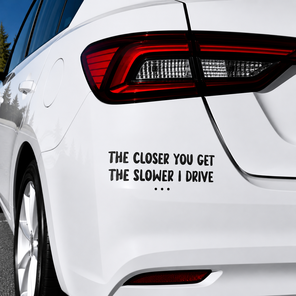 A black vinyl car decal with the humorous text 'THE CLOSER YOU GET THE SLOWER I DRIVE' and three dots, shown applied to the rear left side of a white sedan.