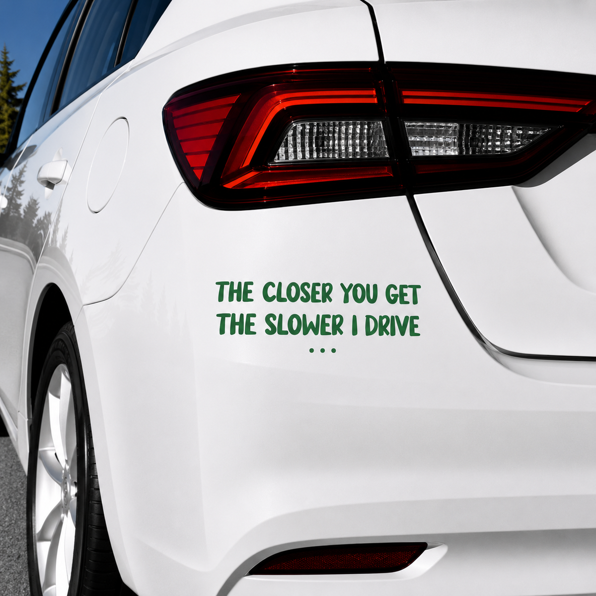 A green text decal on the rear left side of a white car, reading 'THE CLOSER YOU GET THE SLOWER I DRIVE' with three dots underneath, shown outdoors against a blue sky.