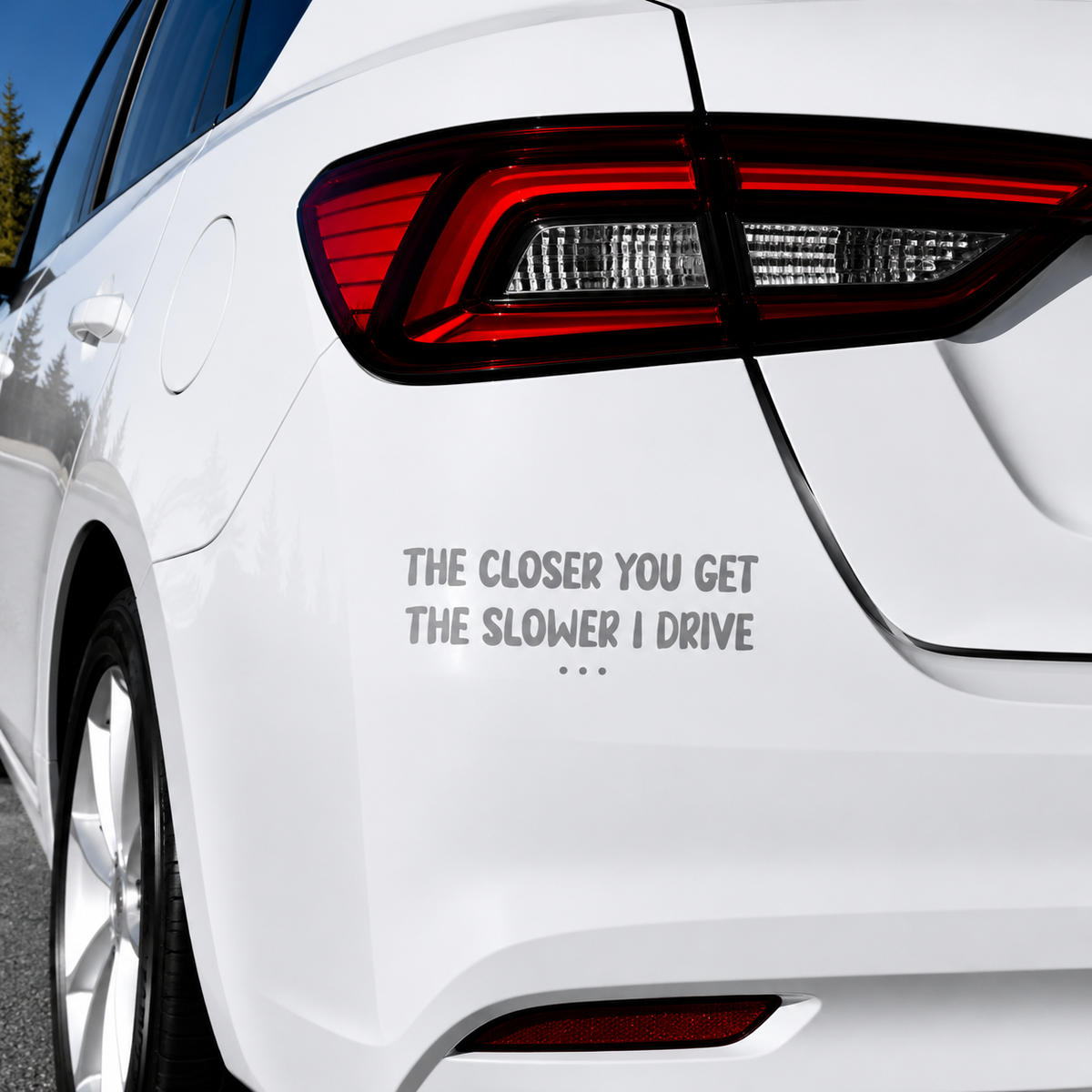 A gray vinyl decal with the text 'THE CLOSER YOU GET THE SLOWER I DRIVE' and three dots, applied to the rear left side of a white car.