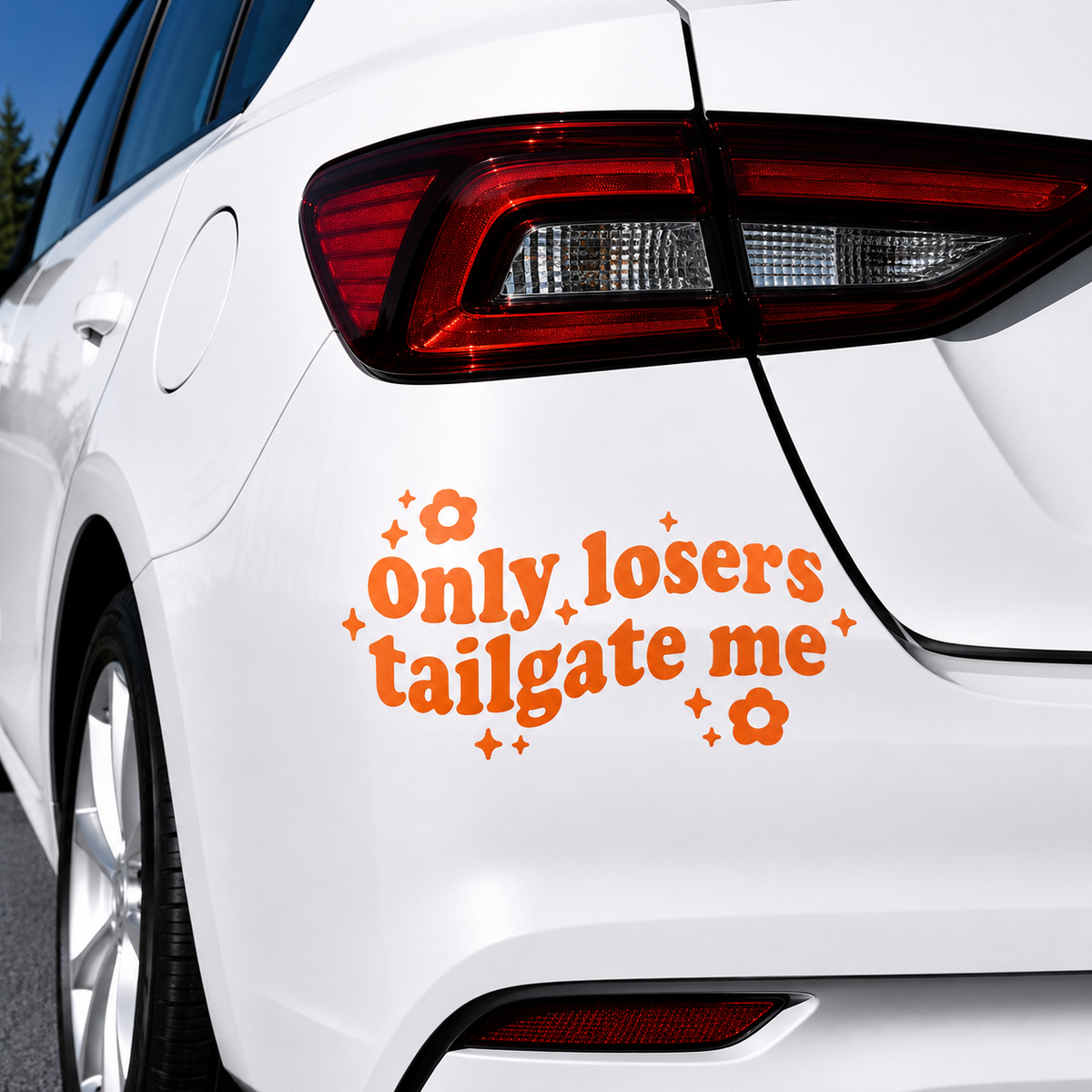An orange vinyl decal with the text 'Only losers tailgate me' and small flower and star accents, applied to the rear quarter panel of a white car.