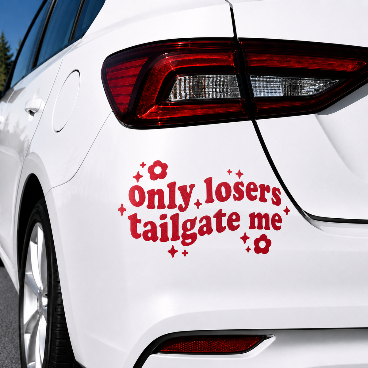 A red vinyl car decal with the text 'Only losers tailgate me' in a retro font, decorated with small red flowers and starburst shapes, shown applied to the rear left side of a white car.