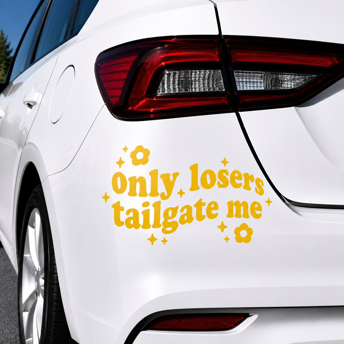 A yellow vinyl decal with the text 'only losers tailgate me' in a retro font, decorated with small yellow flowers and stars, applied to the rear left side of a white car.