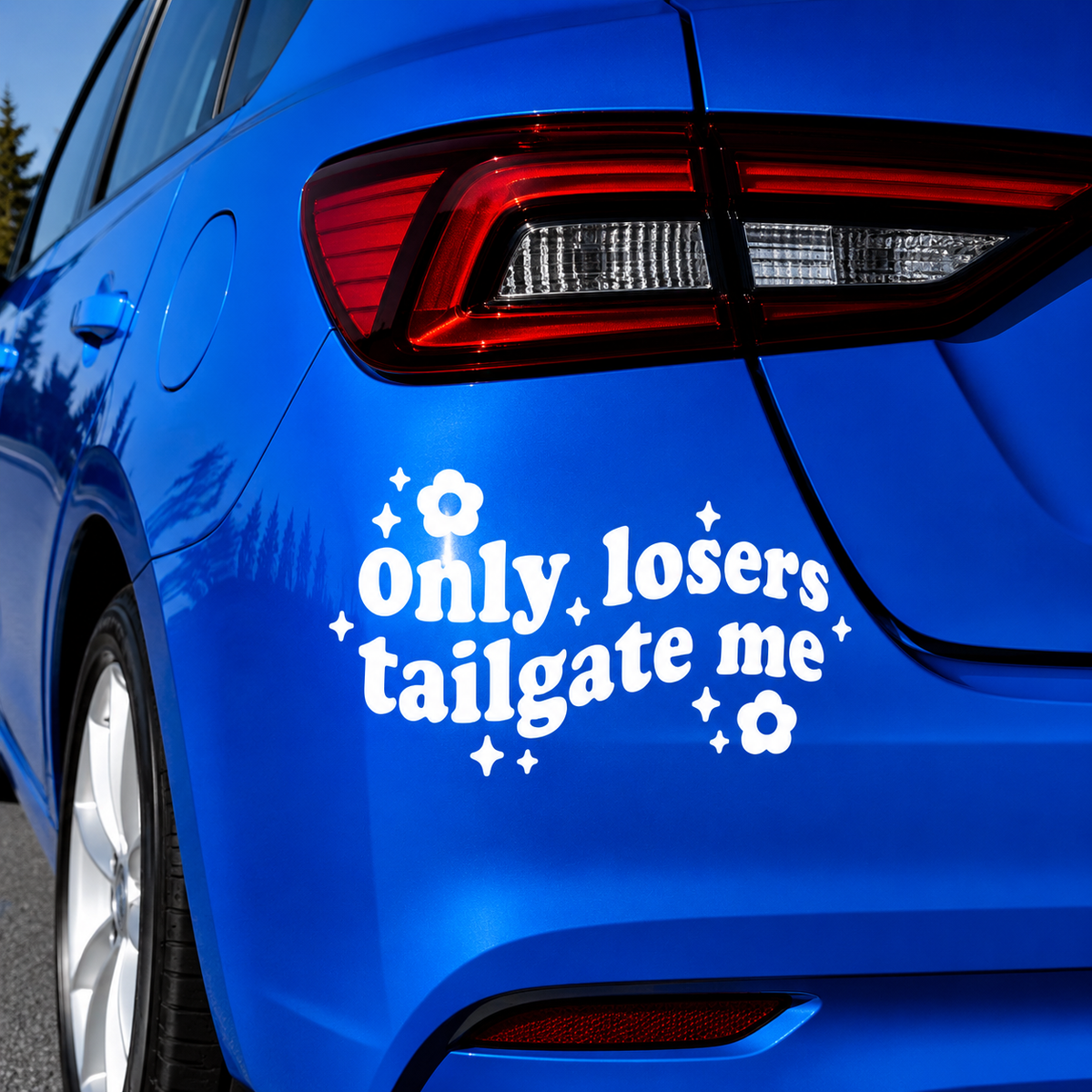 A white vinyl decal with the text 'Only losers tailgate me' surrounded by small white flowers and sparkles, applied to the rear left side of a bright blue car.