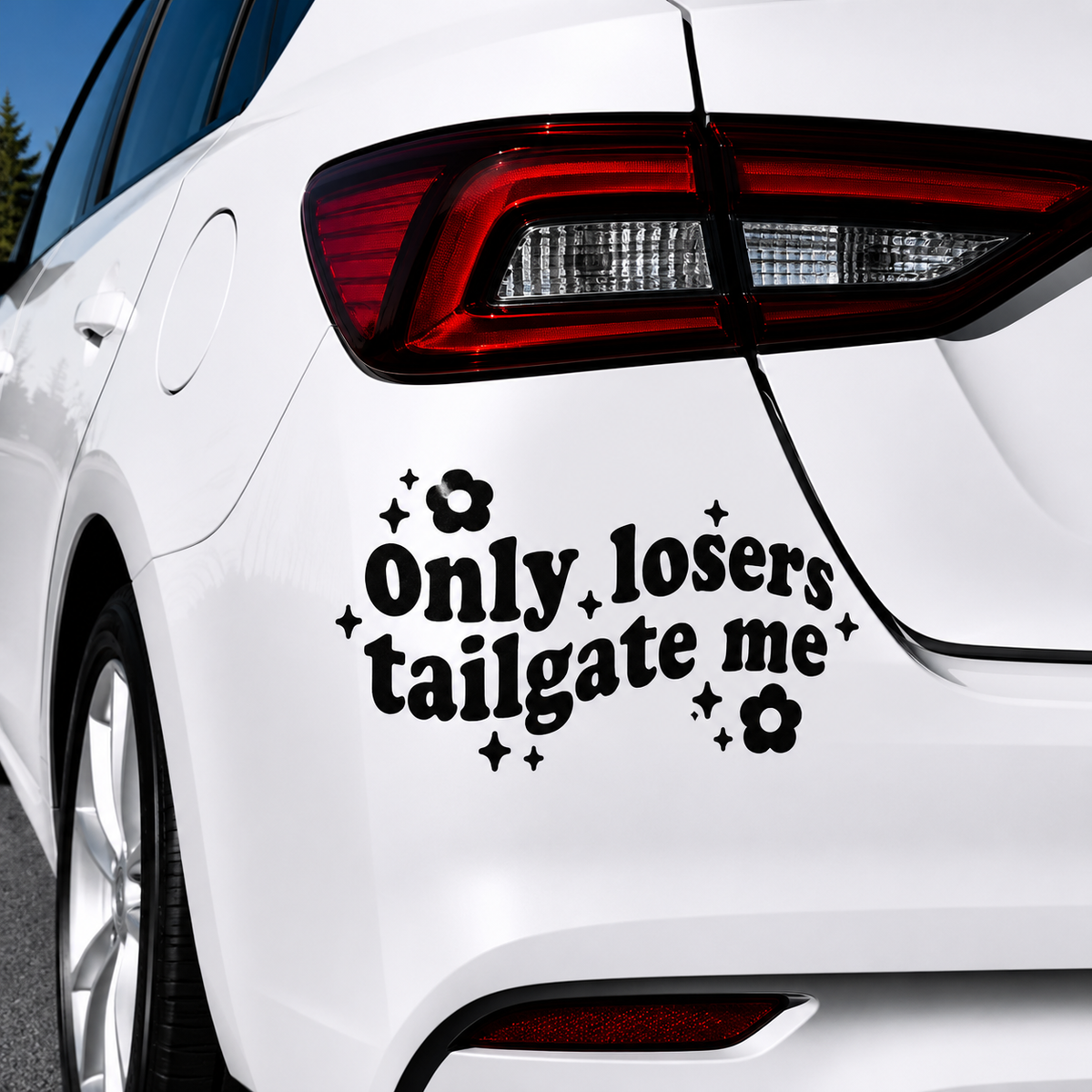 A black vinyl decal with the text 'Only losers tailgate me' in a bold serif font, decorated with small black flowers and sparkles, shown applied to the rear left side of a white car.