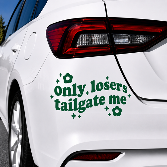 A green vinyl car decal with the text 'only losers tailgate me' and small flower and star accents, shown applied to the rear quarter panel of a white car.