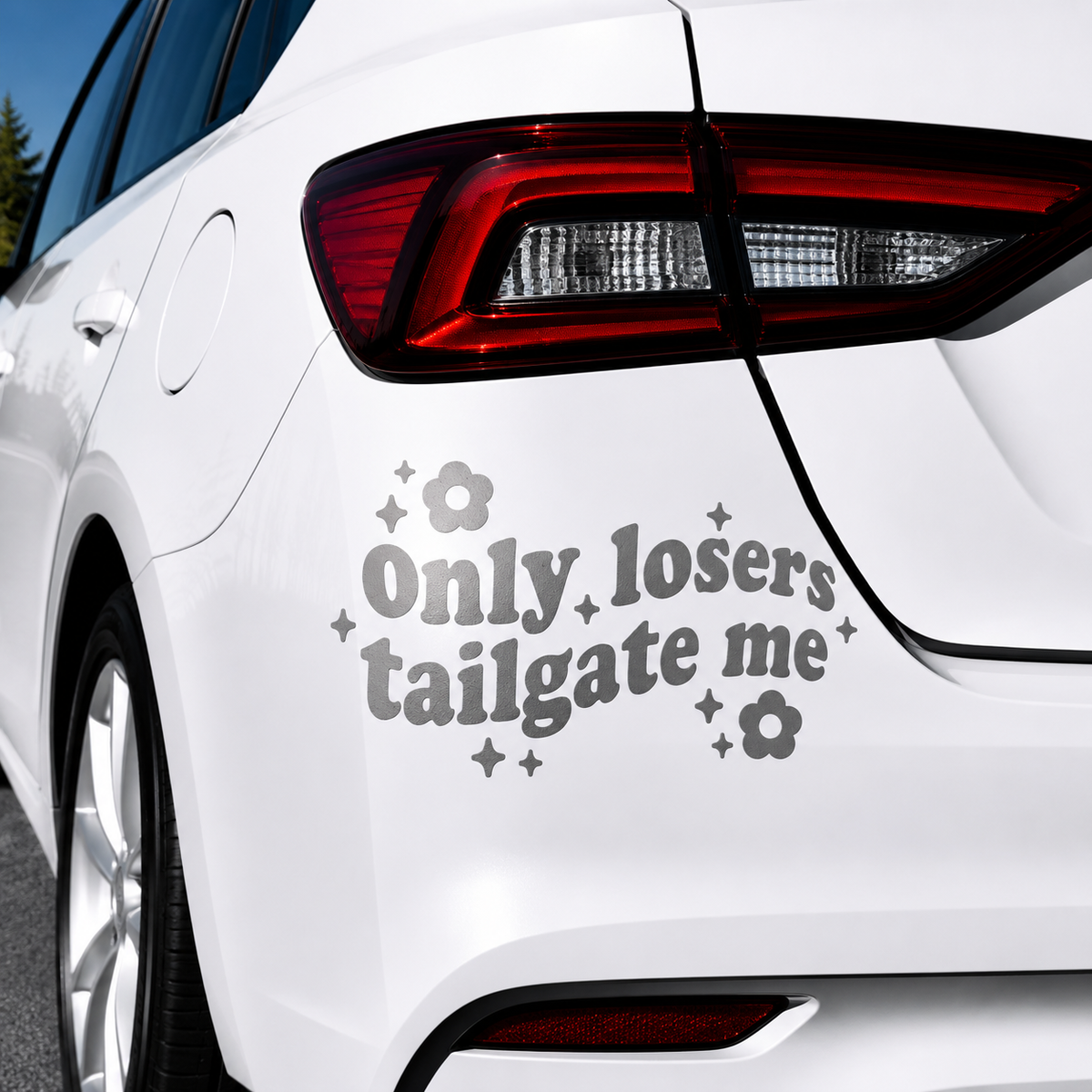 A gray vinyl car decal with the text 'Only losers tailgate me' in a cursive font, decorated with small flower and star motifs, shown applied to the rear quarter panel of a white car.