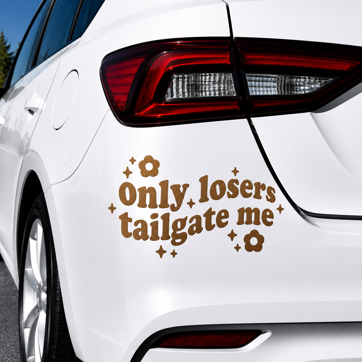 A brown vinyl car decal with the text 'Only losers tailgate me' in a retro font, decorated with small flowers and stars, shown applied to the rear quarter panel of a white car.