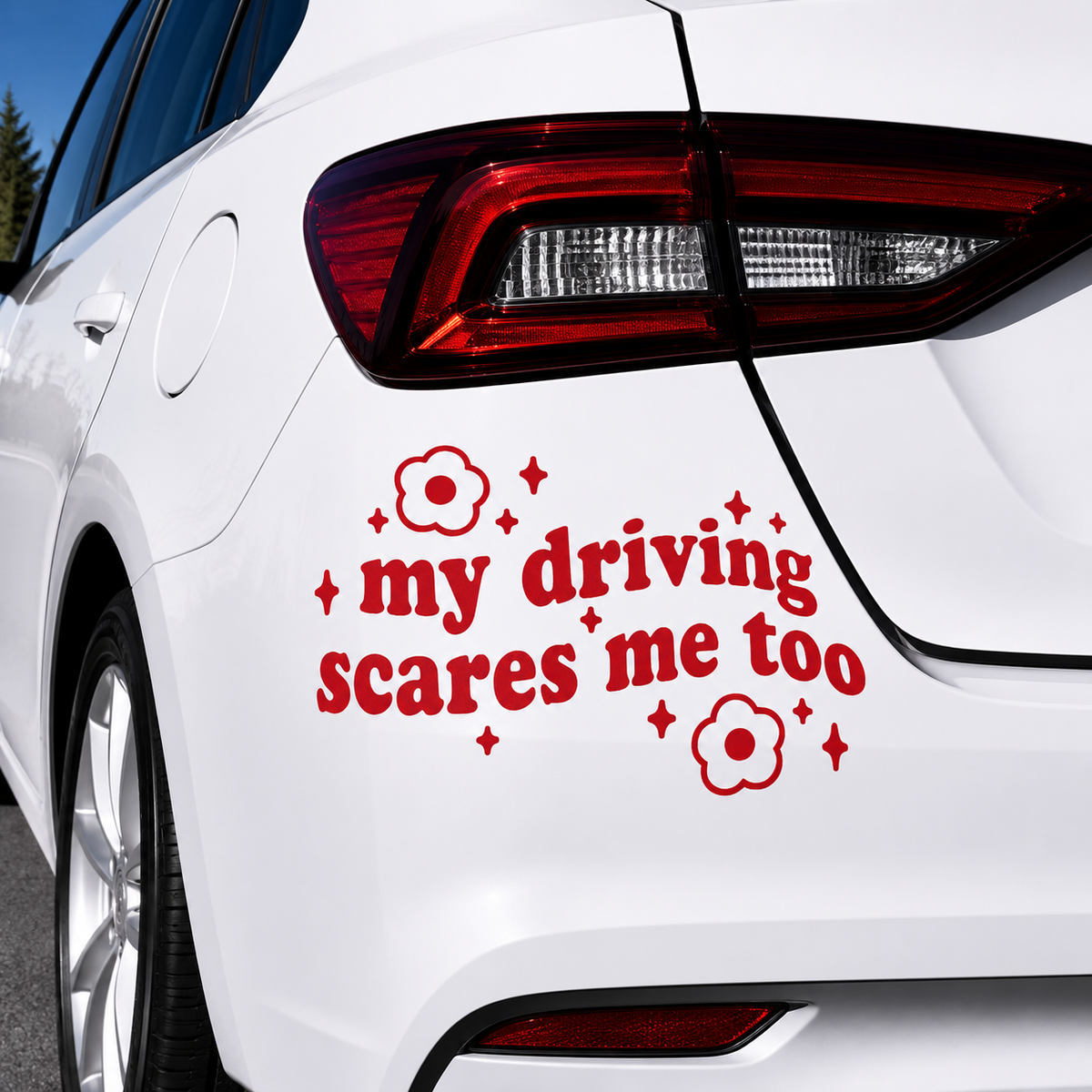 A red vinyl car decal with the text 'my driving scares me too' and small flower and star accents, shown applied to the rear quarter panel of a white car.