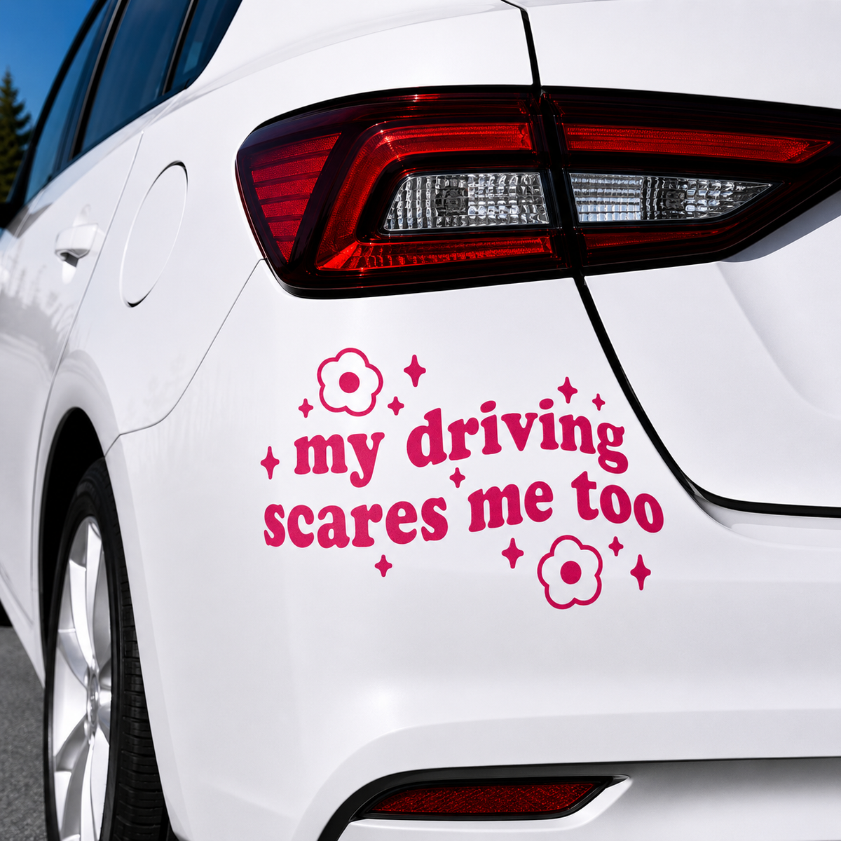 A pink vinyl car decal with the text 'my driving scares me too' and small flower and star accents, shown applied to the rear quarter panel of a white car.