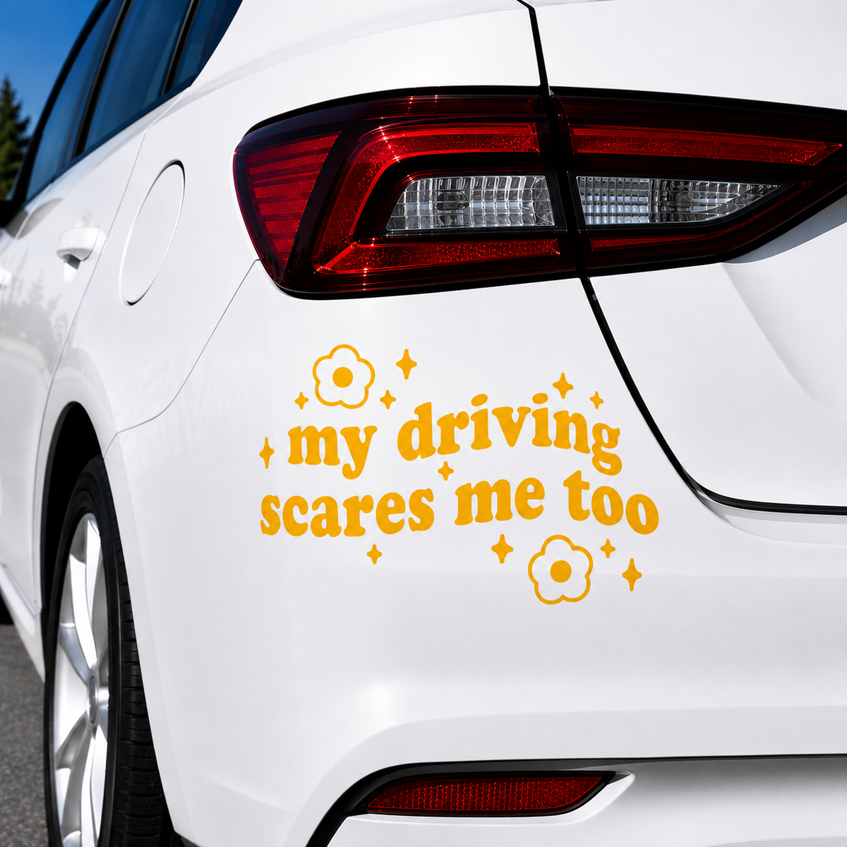 A yellow vinyl car decal with the text 'my driving scares me too' surrounded by small flower and star graphics, applied to the rear quarter panel of a white car.