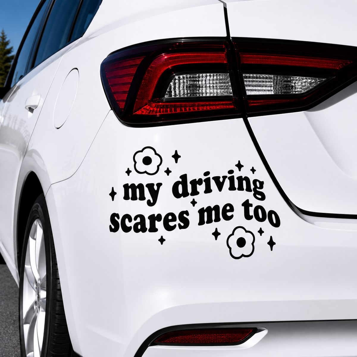 A black vinyl car decal with the text 'my driving scares me too' and small flower and star accents, shown applied to the rear quarter panel of a white car.