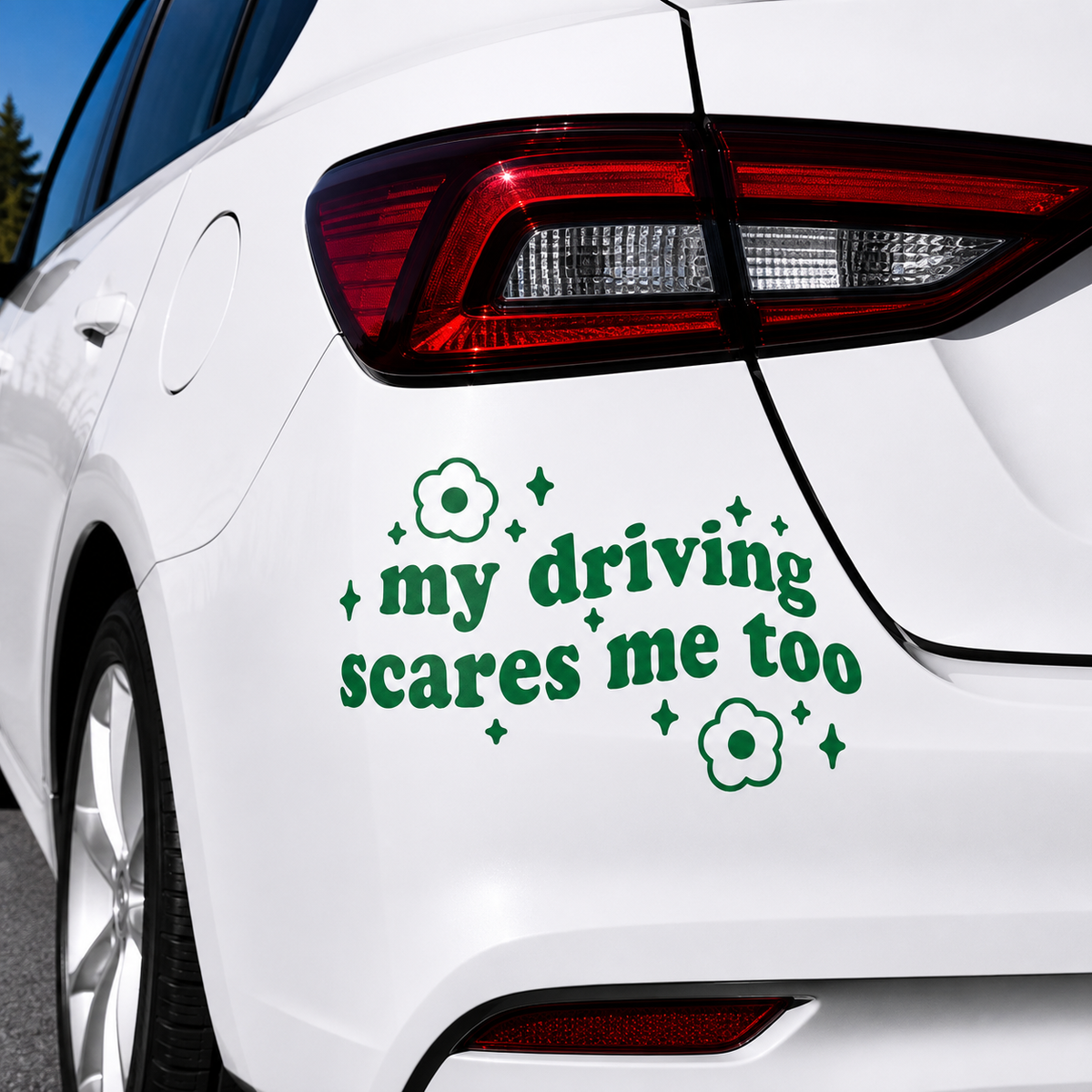A green vinyl car decal with the text 'my driving scares me too' in a playful font, decorated with small flower and star motifs, shown applied to the rear quarter panel of a white car.