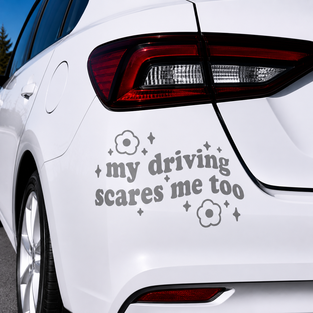 A gray vinyl car decal with the text 'my driving scares me too' and small flower and sparkle graphics, shown applied to the rear quarter panel of a white car.