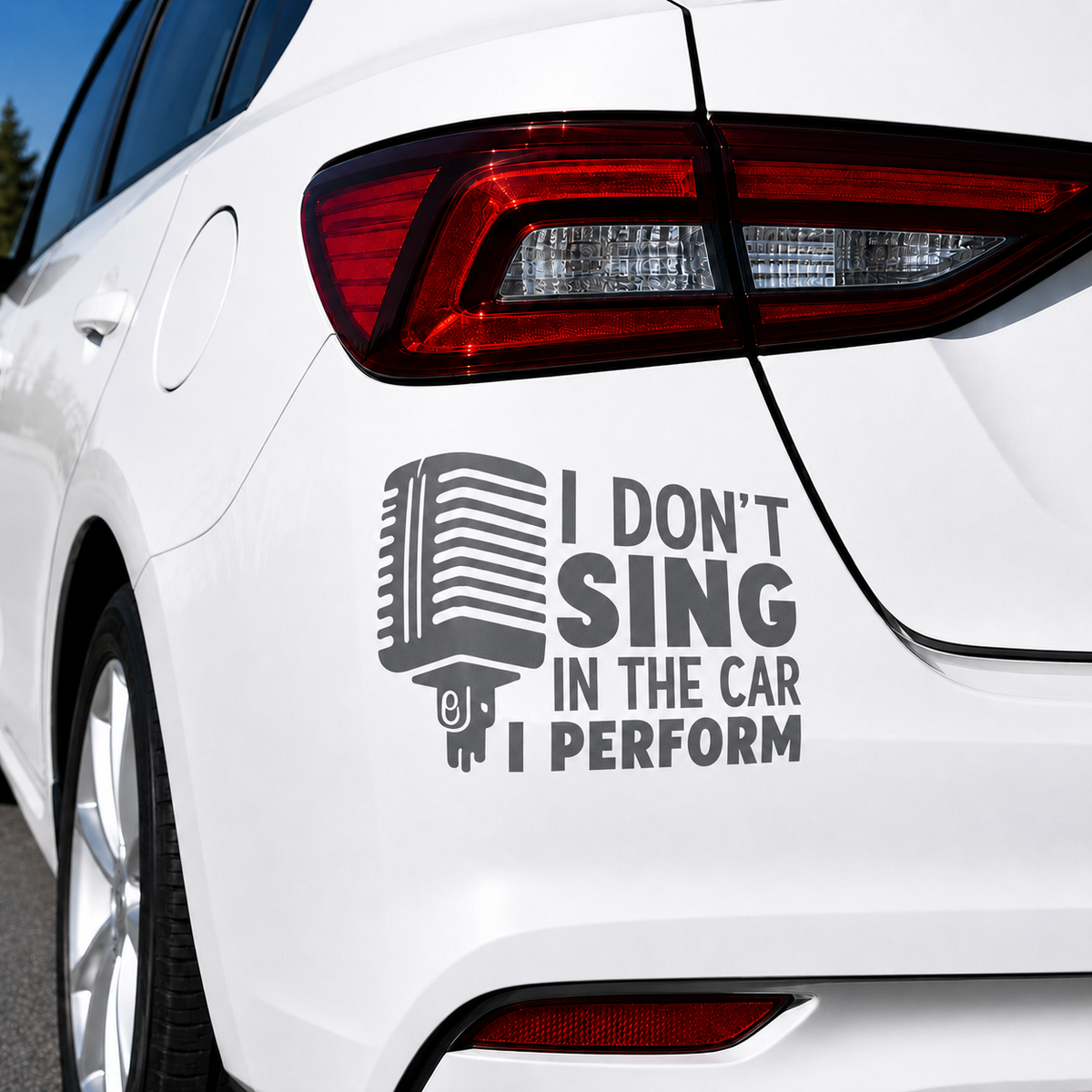 A dark gray vinyl decal applied to the rear quarter panel of a white car. The decal features a retro microphone graphic next to the text 'I DON'T SING IN THE CAR I PERFORM'.