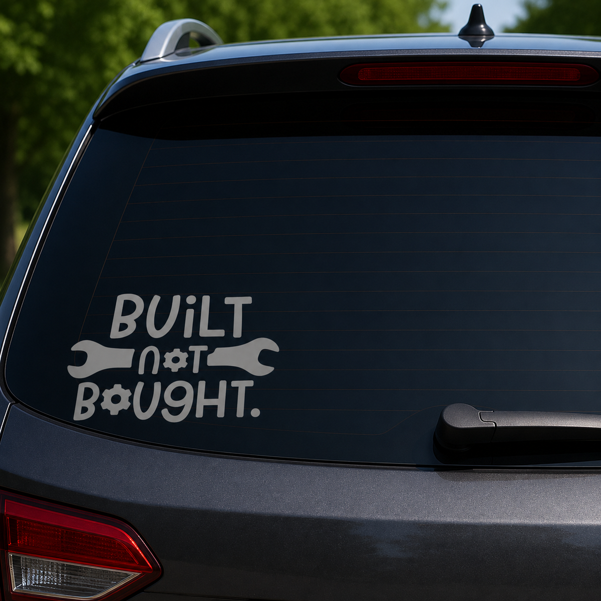 A white vinyl decal on the rear window of a dark gray SUV, featuring the text 'BUILT not BOUGHT.' with wrench and gear icons, set against a blurred green outdoor background.