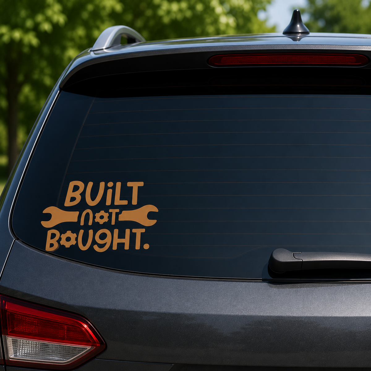 A gold vinyl decal on the rear window of a dark gray SUV, featuring the text 'BUILT not BOUGHT.' with wrench and bolt icons, set against a blurred background of green trees.
