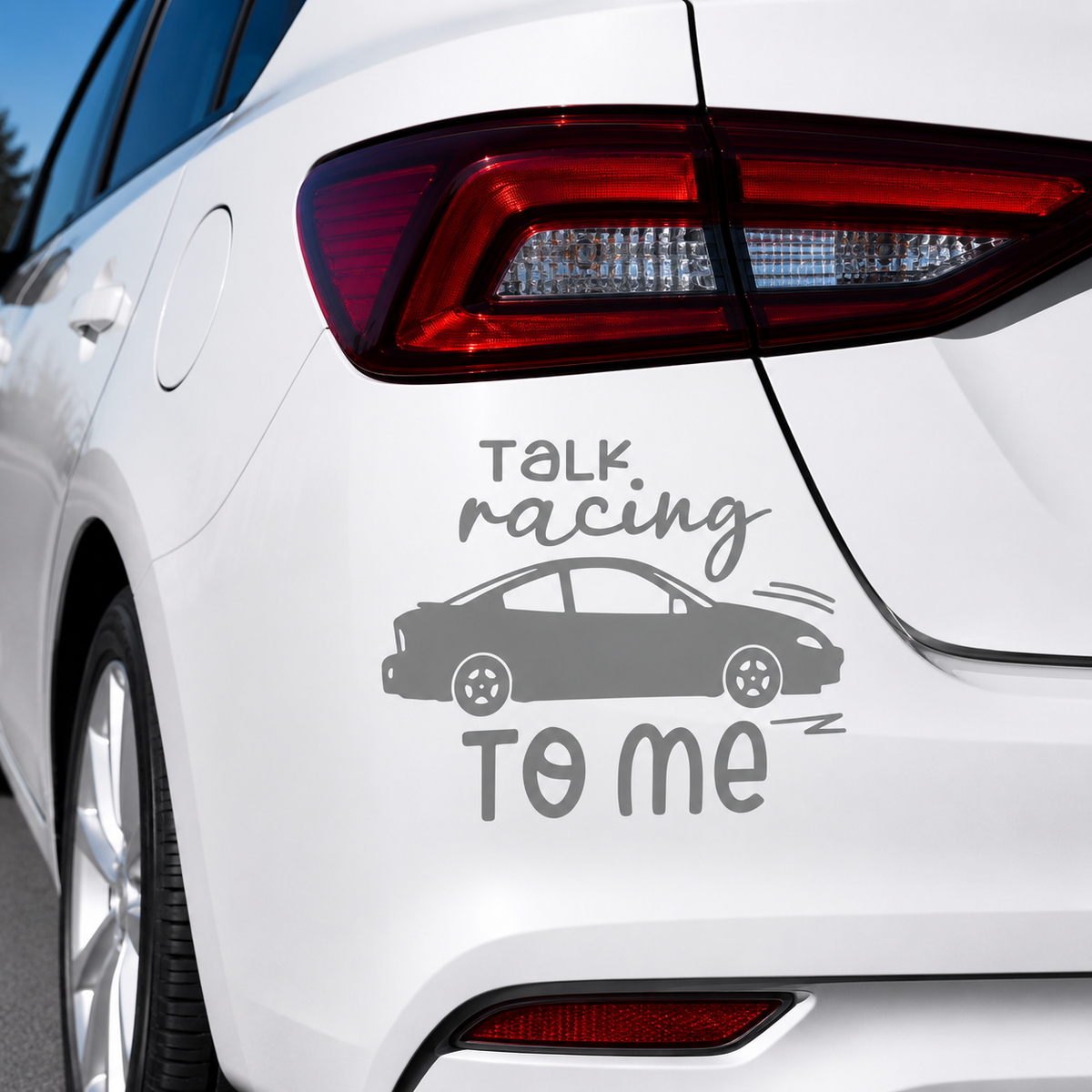 A gray vinyl car decal featuring the text 'TALK racing TO ME' with a simple car silhouette, shown applied to the rear quarter panel of a white car.