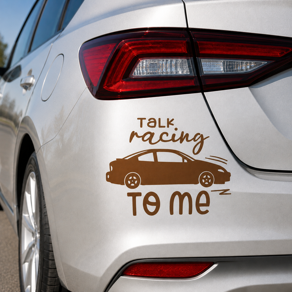 A brown vinyl car decal applied to the rear quarter panel of a silver car. The decal features the text 'TALK racing TO ME' in a stylized font, with a silhouette of a sports sedan in the center.