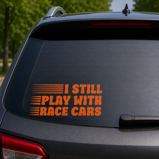An orange vinyl decal with the text 'I STILL PLAY WITH RACE CARS' and horizontal racing stripes, shown applied to the back window of a dark gray SUV.