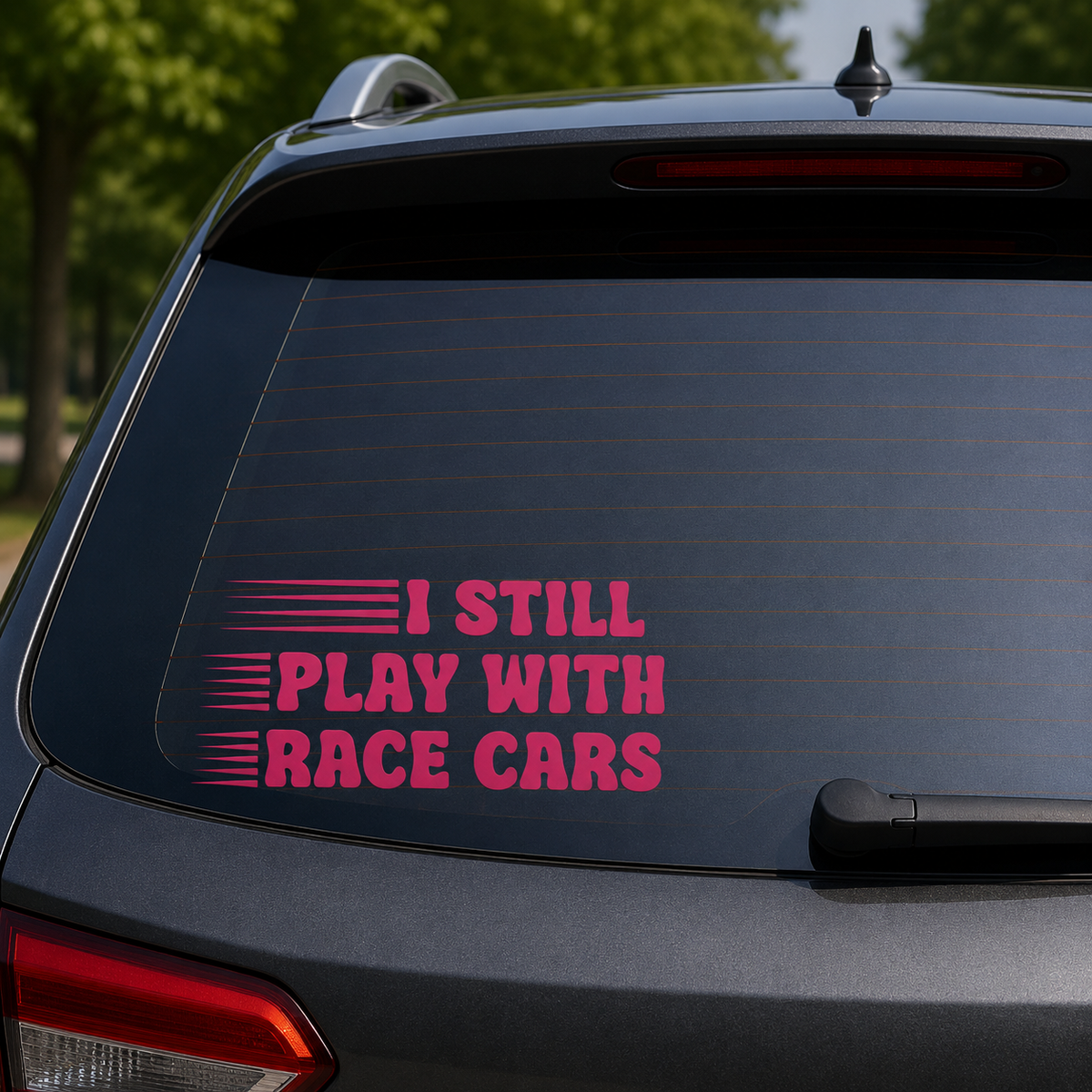 A pink vinyl decal with the text 'I STILL PLAY WITH RACE CARS' and horizontal stripe graphics, applied to the rear window of a dark gray SUV. The background shows blurred green trees outdoors.