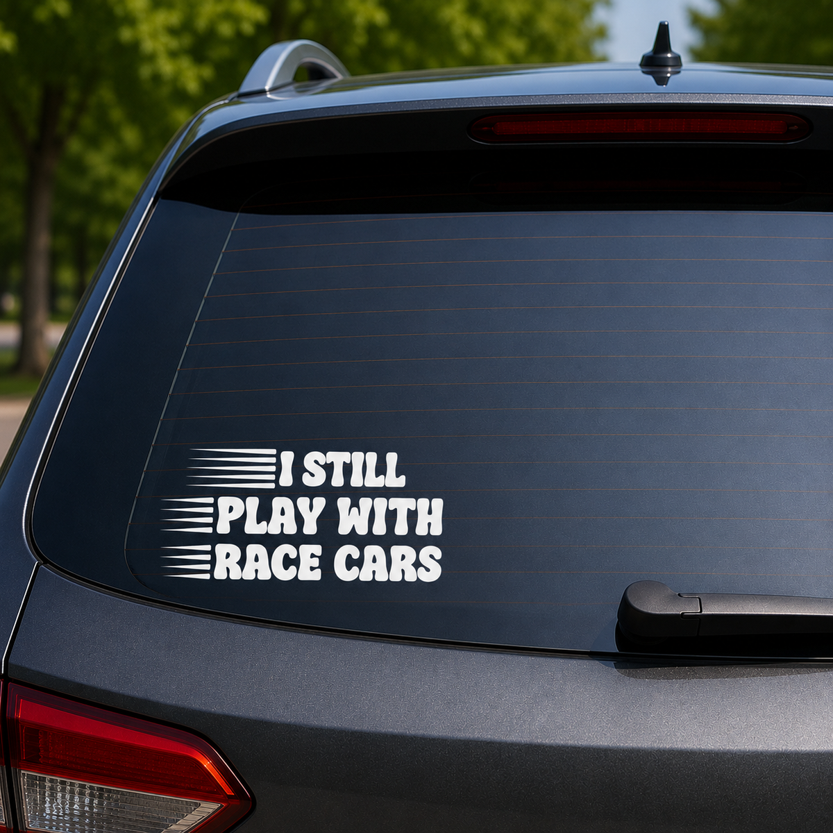 A white vinyl decal with the text 'I STILL PLAY WITH RACE CARS' and horizontal racing stripes, applied to the back window of a dark gray SUV.