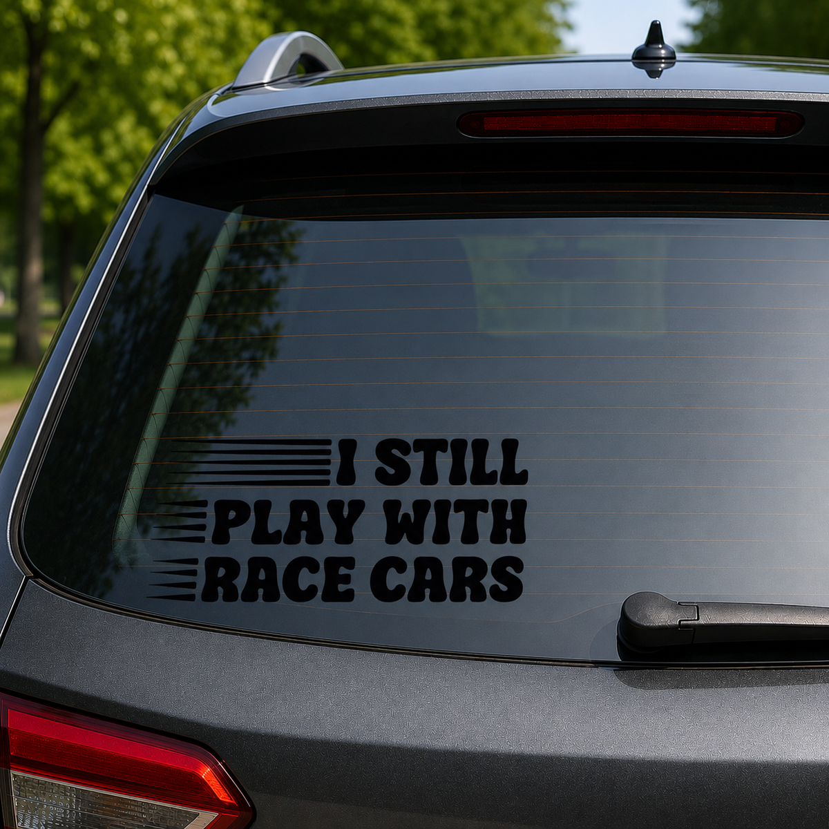 A black vinyl decal with the text 'I STILL PLAY WITH RACE CARS' and horizontal stripe graphics, applied to the rear window of a dark gray SUV. The background shows blurred green trees and outdoor scenery.