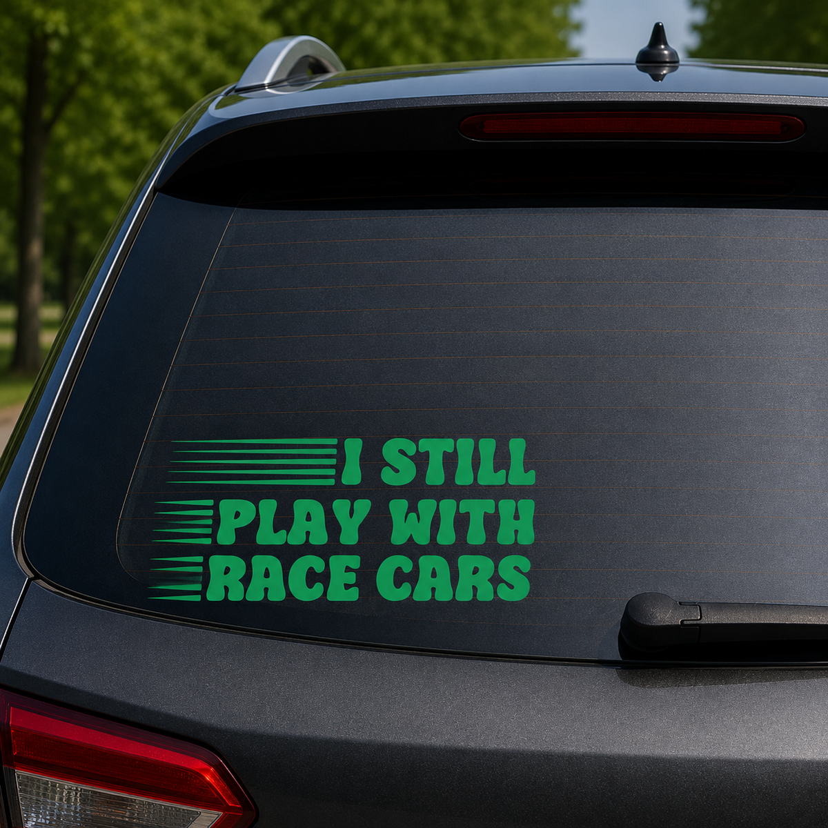 A green vinyl decal with the text 'I STILL PLAY WITH RACE CARS' applied to the rear window of a dark gray SUV, with blurred green trees in the background.