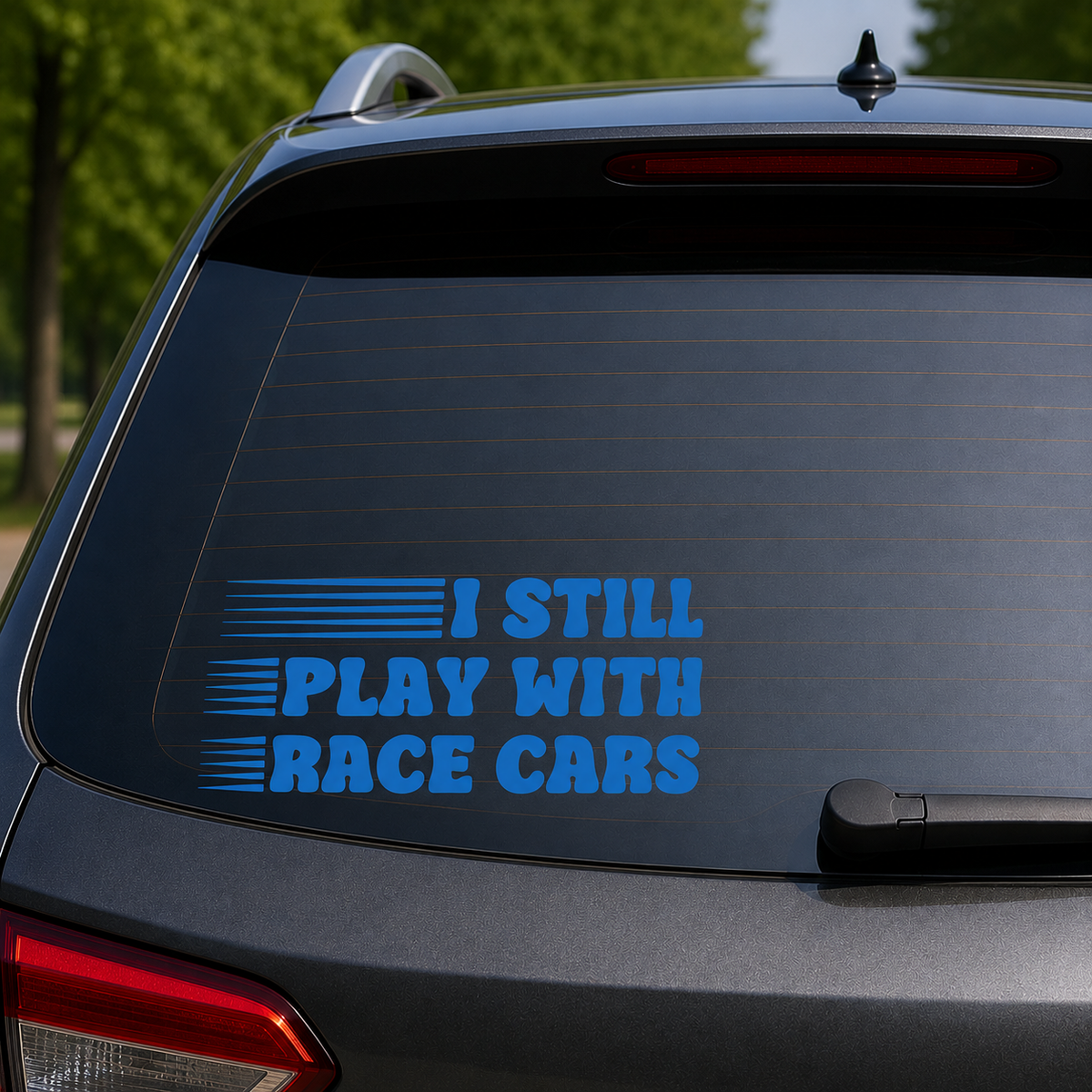 A blue vinyl decal reading 'I STILL PLAY WITH RACE CARS' applied to the rear window of a dark gray SUV, with blurred green trees in the background.