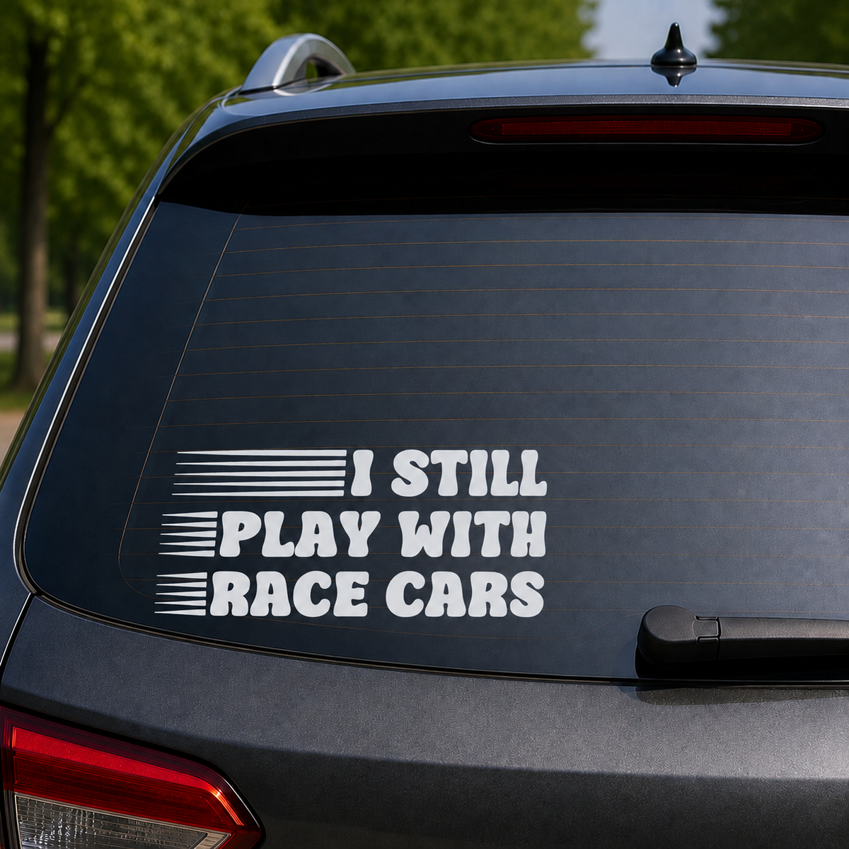 A white vinyl decal with the text 'I STILL PLAY WITH RACE CARS' and horizontal stripe graphics, applied to the rear window of a dark gray SUV.