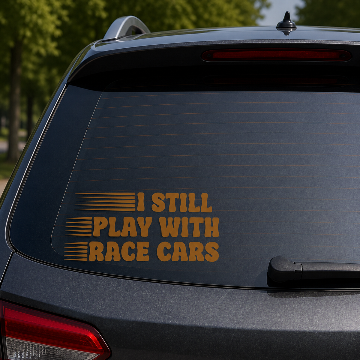 A gold vinyl decal reading 'I STILL PLAY WITH RACE CARS' with horizontal stripe accents, shown applied to the rear window of a dark gray SUV.