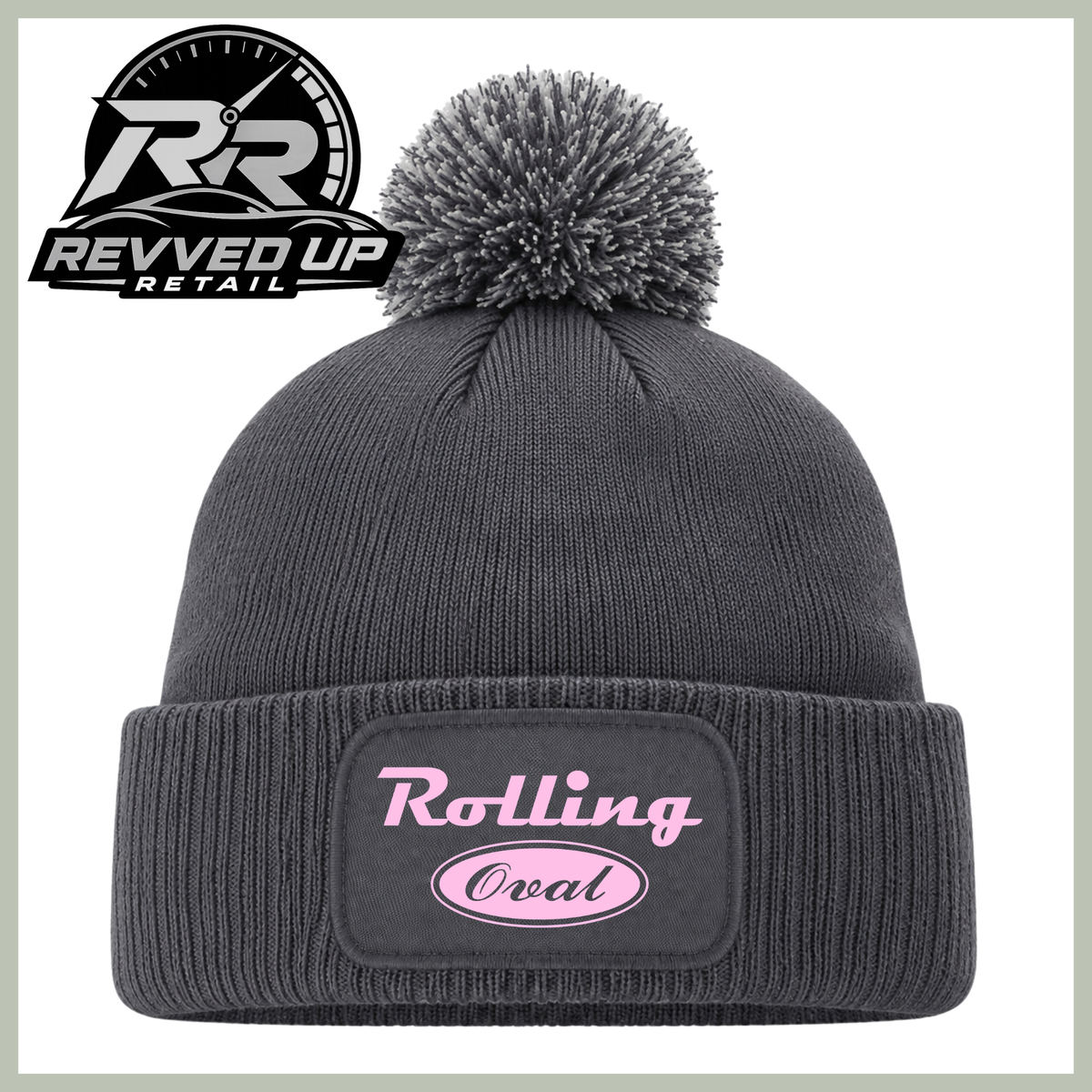 A dark gray ribbed knit pom-pom beanie with a cuffed brim featuring a black patch with pink text that reads 'Rolling Oval'. The Revved Up Retail logo is displayed in the top-left corner.