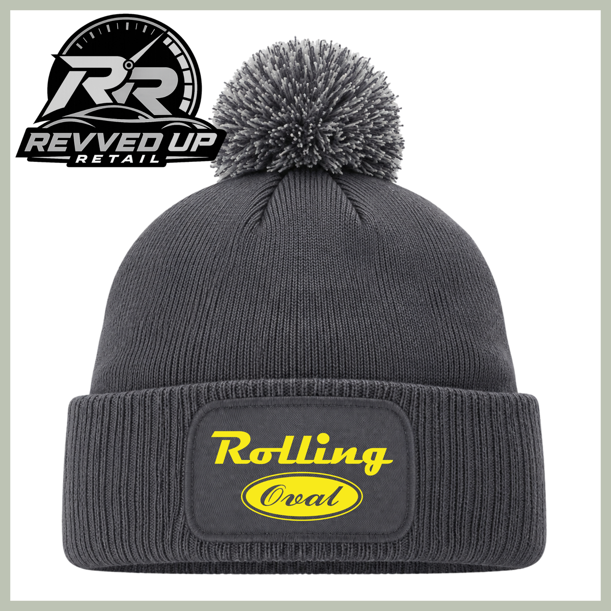 A dark gray ribbed knit pom-pom beanie with a cuffed brim, featuring a yellow 'Rolling oval' logo patch on the front. The image includes a Revved Up Retail logo overlay in the top-left corner.