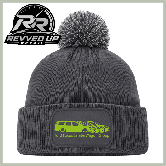 A dark gray ribbed knit beanie with a matching pom-pom and a folded cuff featuring a rectangular patch with a bright green car graphic and the text 'Ford Focus Estate/Wagon Group.' A Revved Up Retail logo appears in the top-left corner.