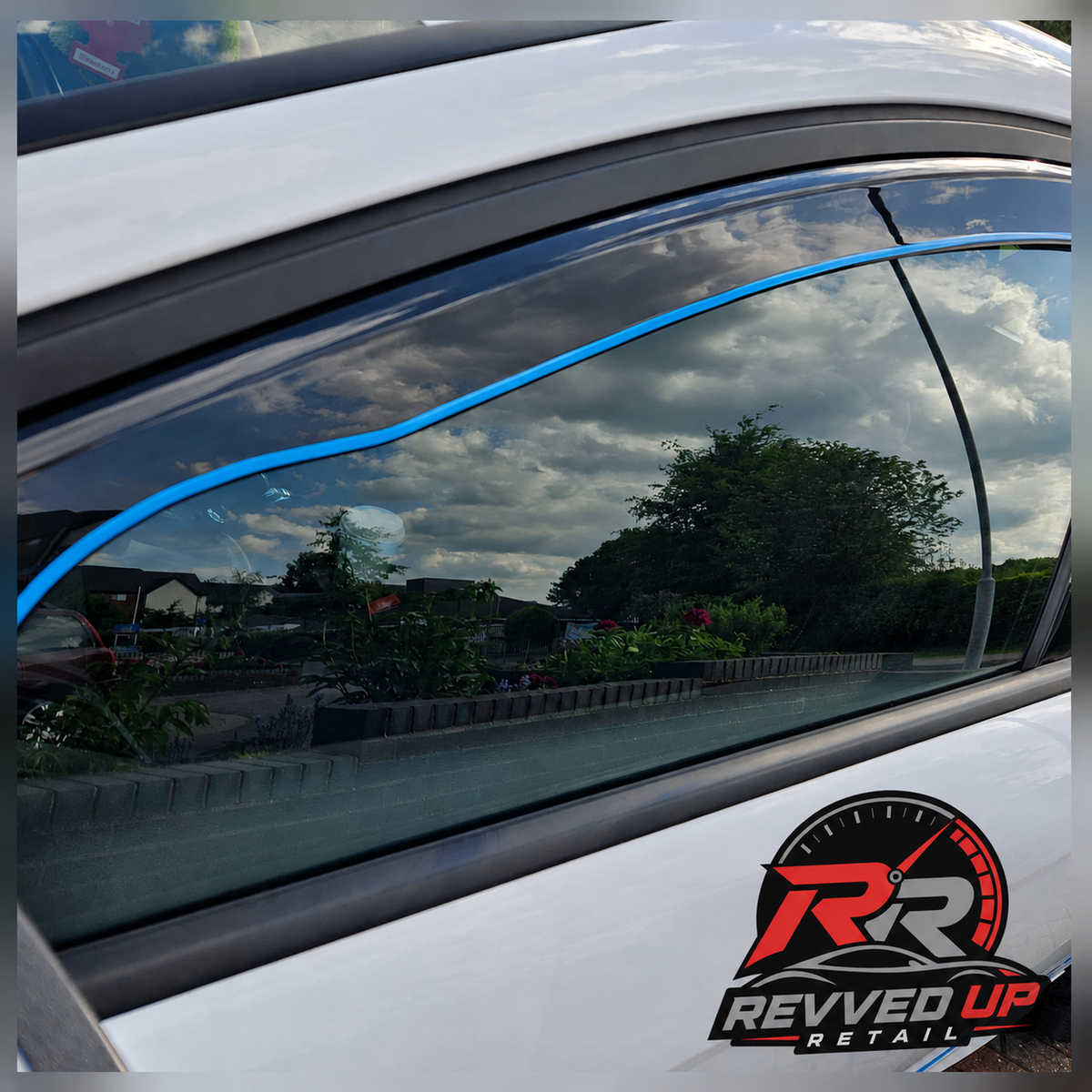 A close-up of a white car door window showing a bright blue tinted film applied along the upper window frame, with a Revved Up Retail logo sticker visible in the bottom right corner.
