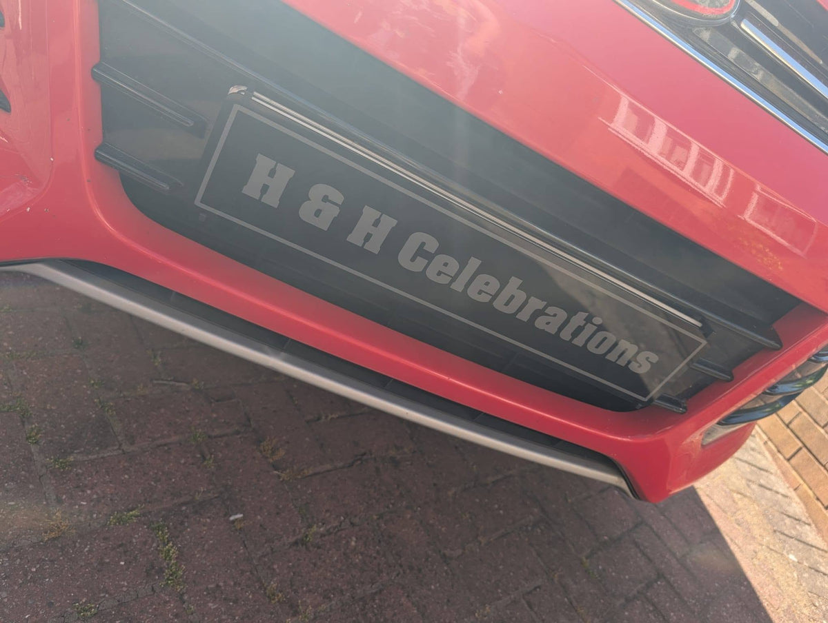 A close-up of a red car's front bumper area, showing a black grille insert with the text 'H & H Celebrations' in white. The car is parked on a paved surface.
